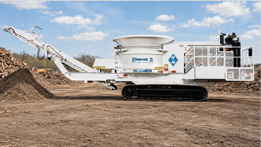 Worker in a hard hat and high-visibility vest stands beside a Diamond Z 1245BTK tracked wood grinder at an outdoor industrial site.