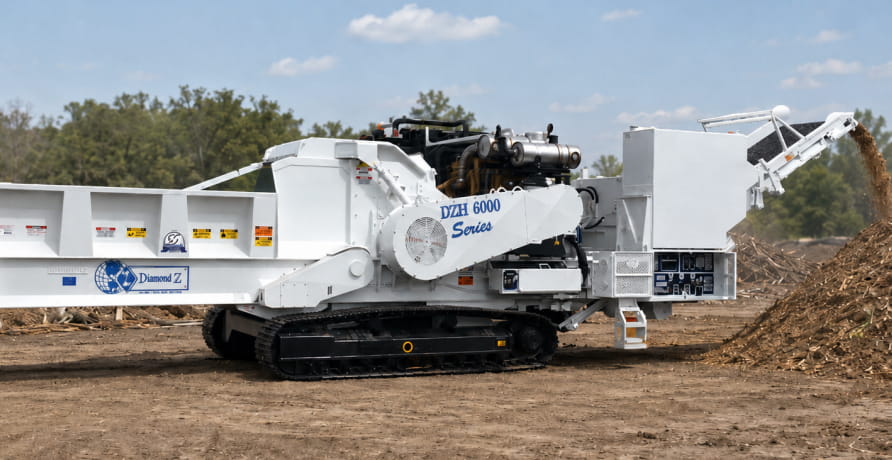 A white Diamond Z track-mounted grinder discharging wood chips or mulch onto a ground pile in a wooded outdoor setting.
