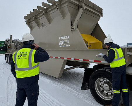 Two workers in hard hats attending to a light-brown industrial hopper on a trailer in snowy conditions.