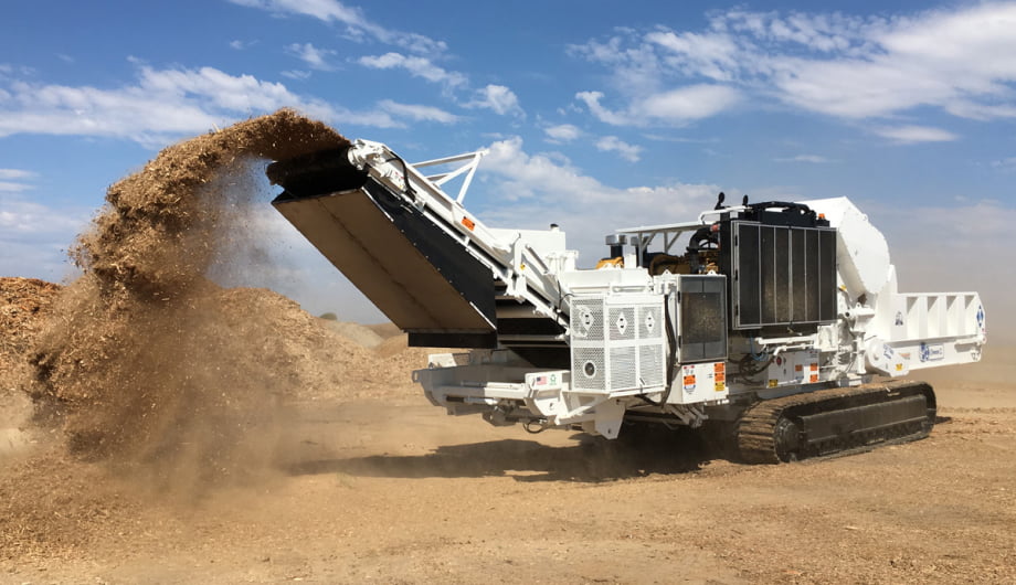 A large white tracked industrial grinder discharges wood chips onto a pile under a blue sky.